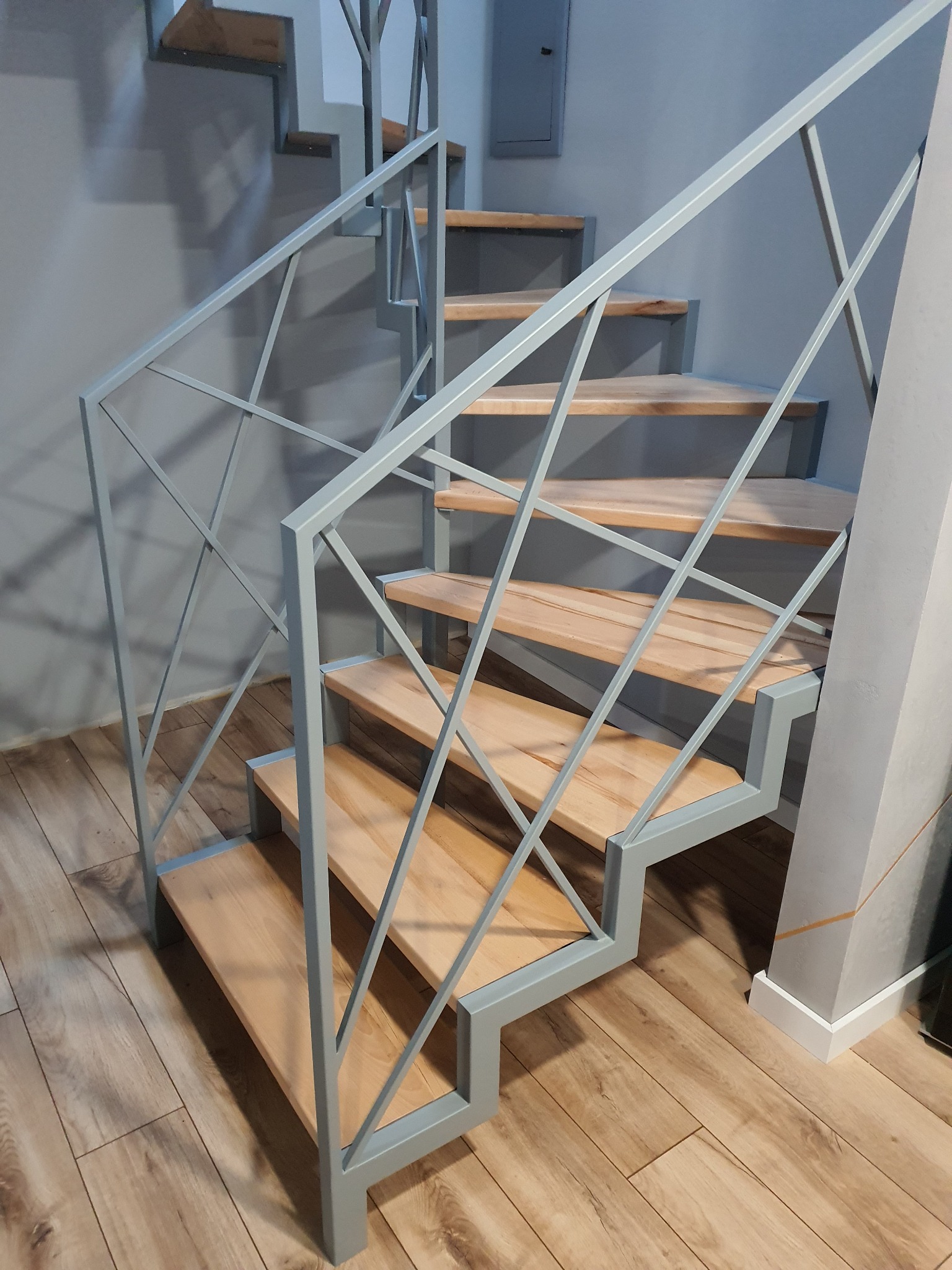 Schody drewniane z metalową, geometryczną balustradą w odcieniach szarości, widoczne z dołu, na tle szarej ściany i drewnianej podłogi.