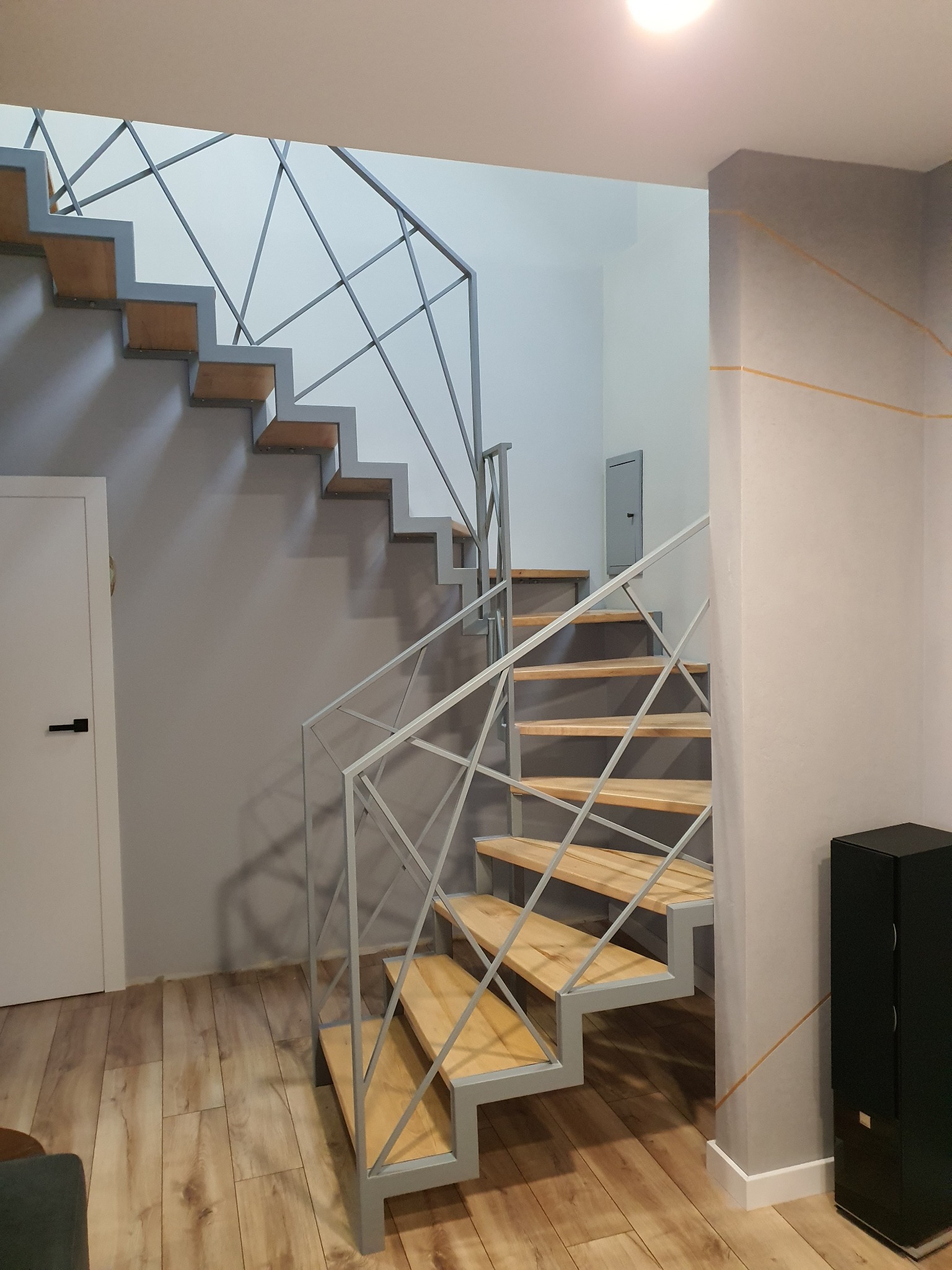 Nowoczesne schody z drewnianymi stopniami i metalową, geometryczną balustradą w szaro-białej tonacji, widok z dołu, z fragmentem drzwi po lewej stronie i kolumną po prawej.
