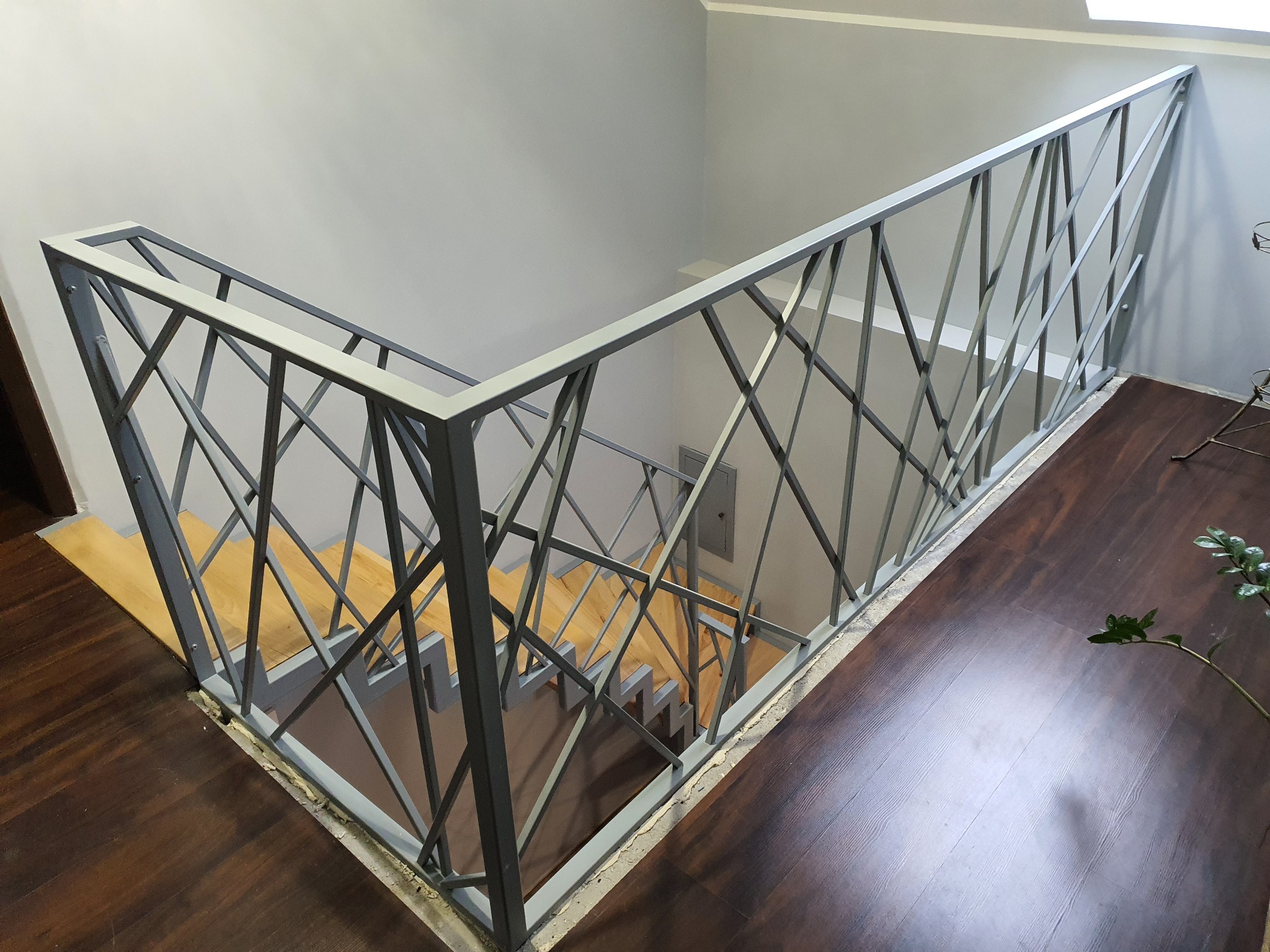 Metalowa balustrada schodowa z geometrycznym wzorem, widok z góry na drewniane schody i ciemną podłogę.