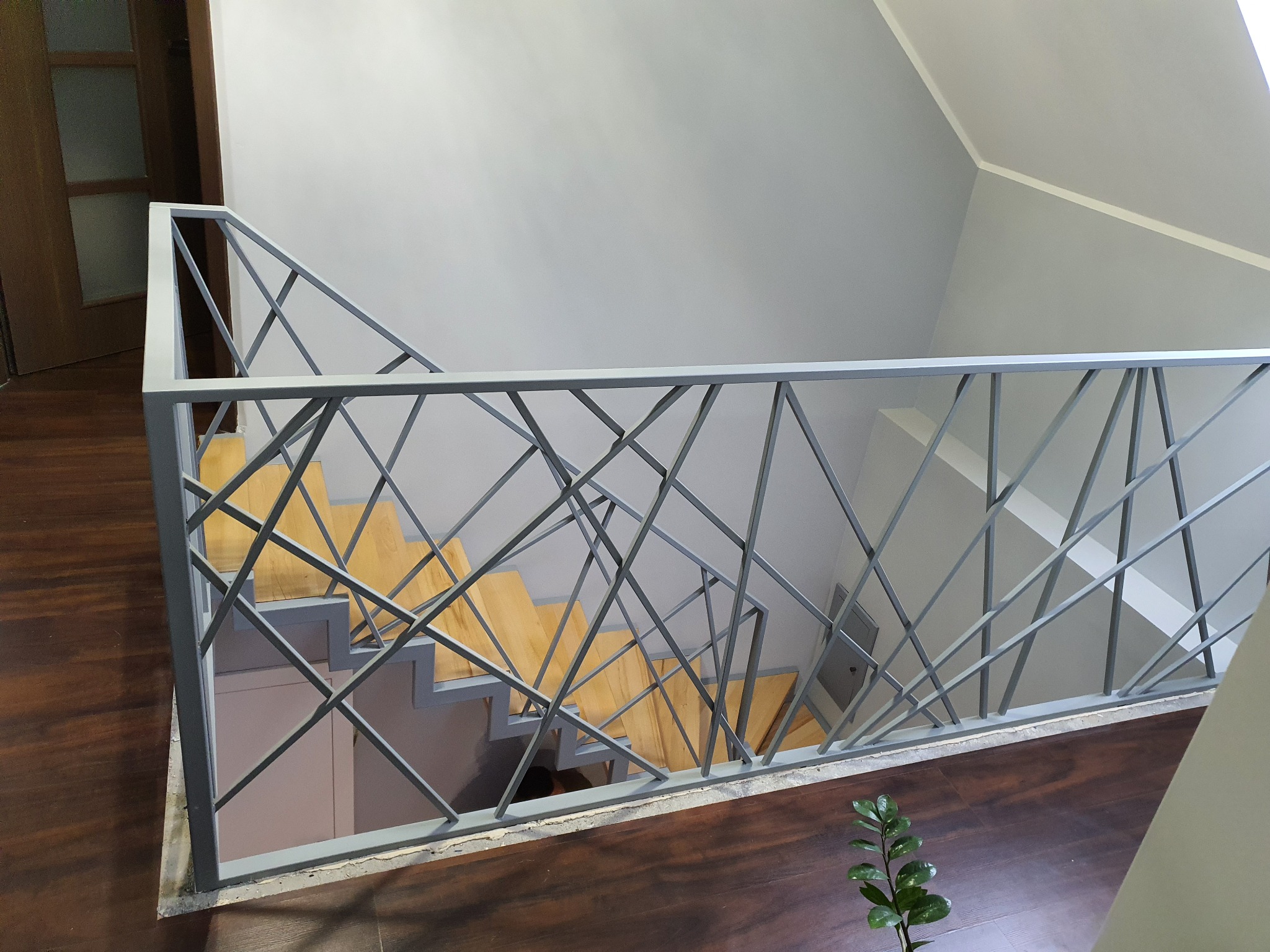Schody drewniane z nowoczesną, geometryczną balustradą ze stali malowanej proszkowo na szaro, widok z góry na ciemną podłogę i rosnącą roślinę doniczkową.