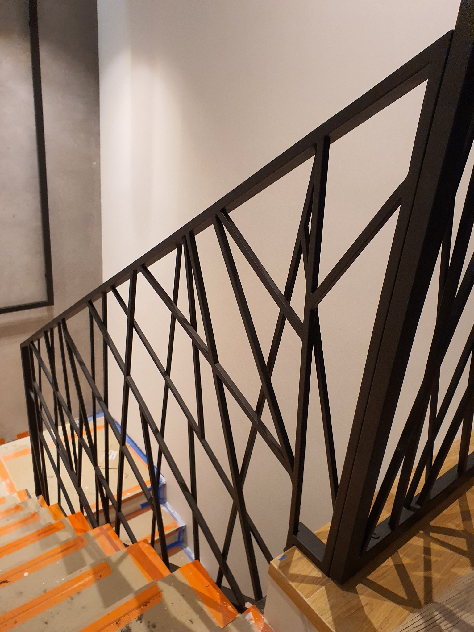 Nowoczesna, czarna balustrada schodowa o geometrycznym wzorze na tle jasnej ściany i zabezpieczonych schodów.