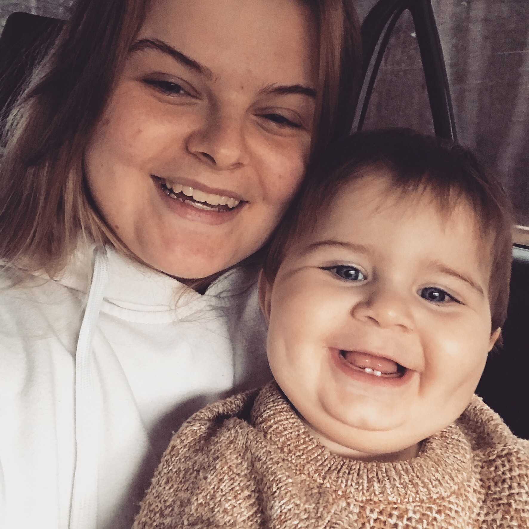 Uśmiechnięta niania w białej bluzie z kapturem, robiąca selfie z radosnym niemowlęciem w wełnianym sweterku, siedząc w samochodzie.