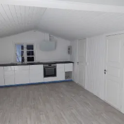 Jasne wnętrze z białymi, panelowymi ścianami i sufitem, czarnym blatem kuchennym i jasną podłogą. Widoczne białe szafki kuchenne, okno, dwie białe drzwi i czarna torba na podłodze.