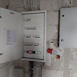 Jagtech Radosław Bagiński - Otwarta skrzynka elektryczna z bezpiecznikami na surowej ścianie. Widoczne kable, gniazdo siłowe i druga, zamknięta skrzynka. Etap prac instalacyjnych.