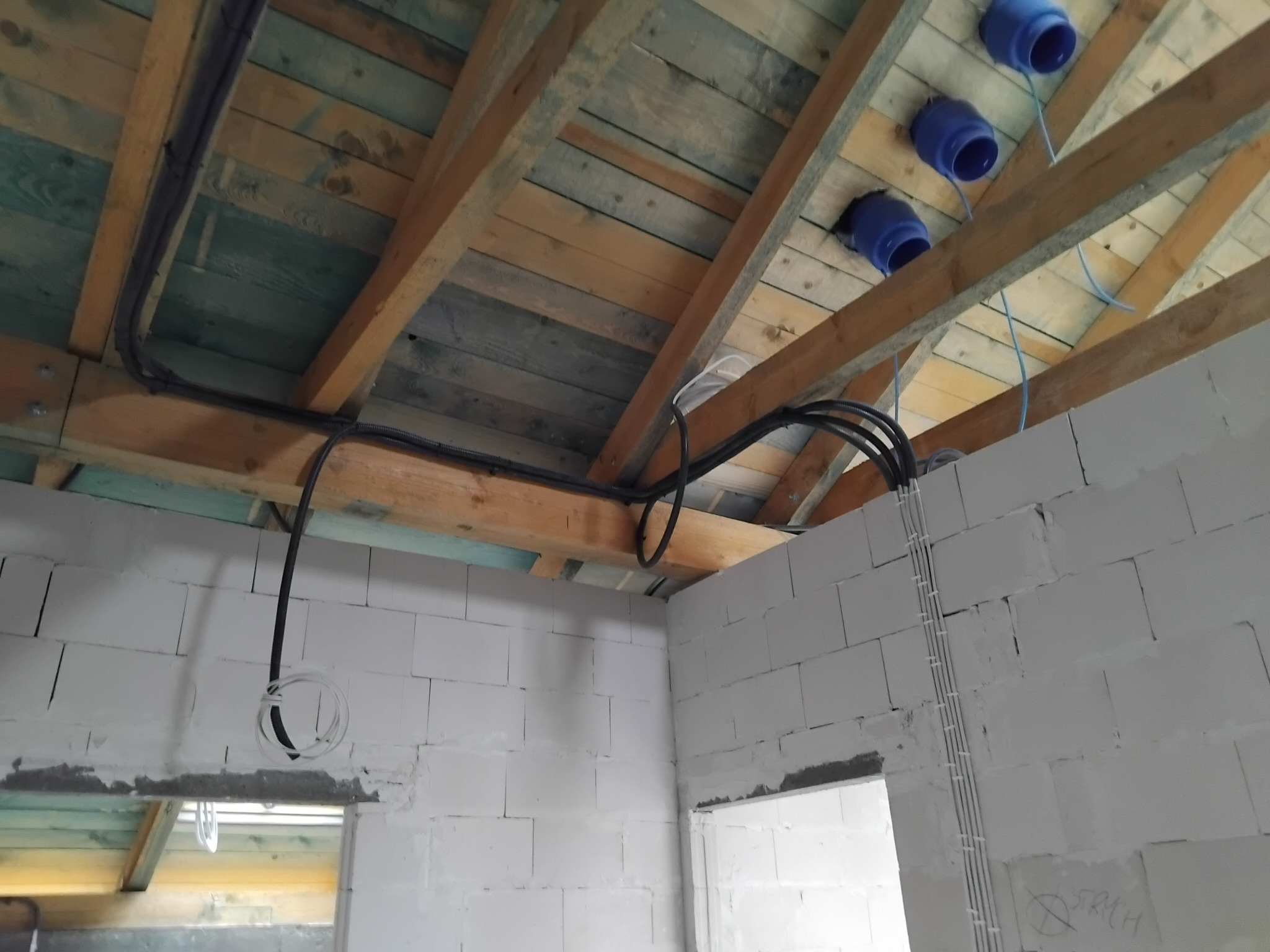 Wnętrze w trakcie budowy z widoczną instalacją elektryczną: czarne kable poprowadzone po drewnianej konstrukcji dachu i białe przewody w ścianie z bloczków.