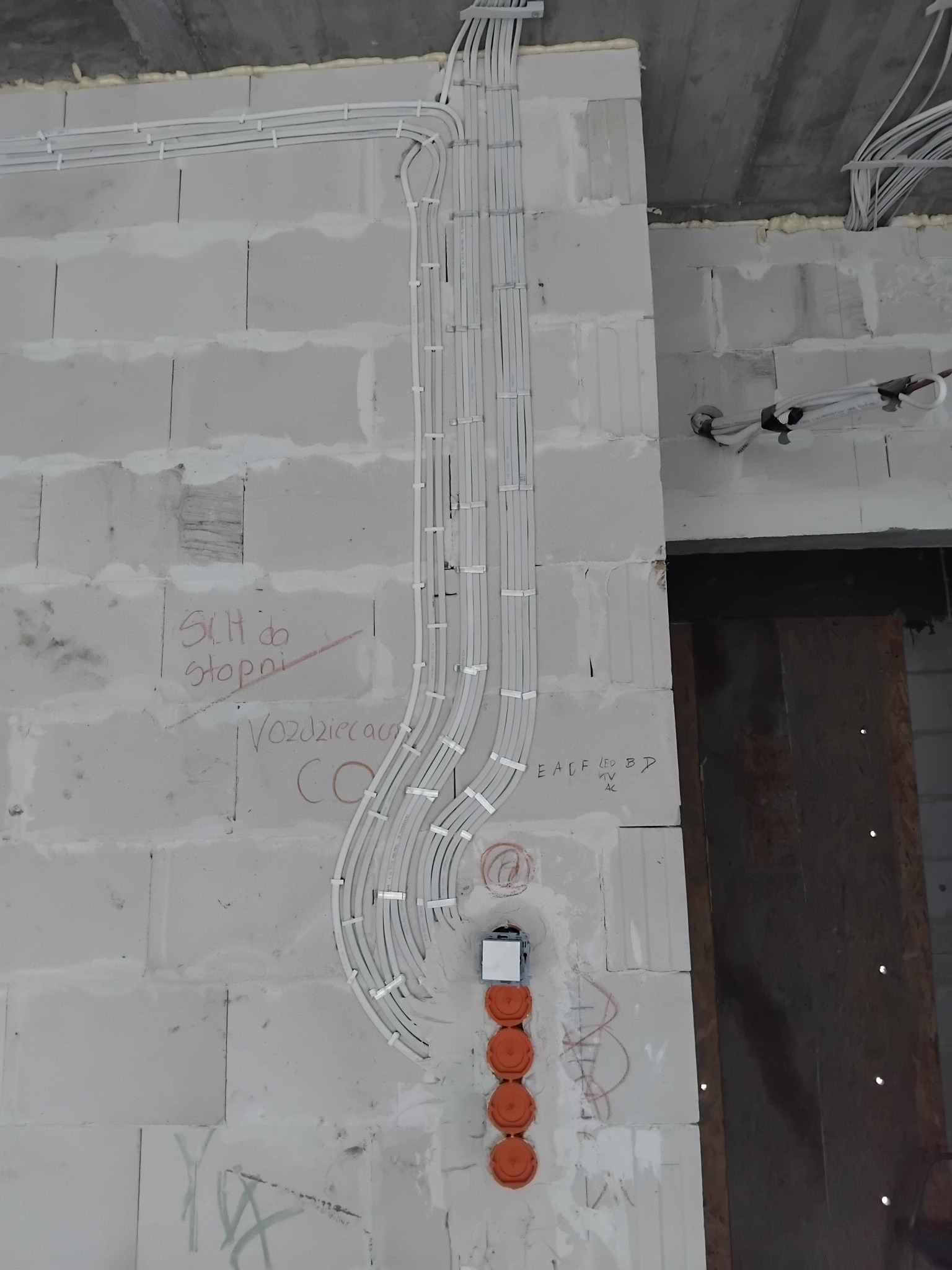 Białe kable elektryczne prowadzone po ścianie z bloczków w Czeczewie, z zamontowanymi puszkami instalacyjnymi. Widoczne napisy na ścianie i fragment sufitu.