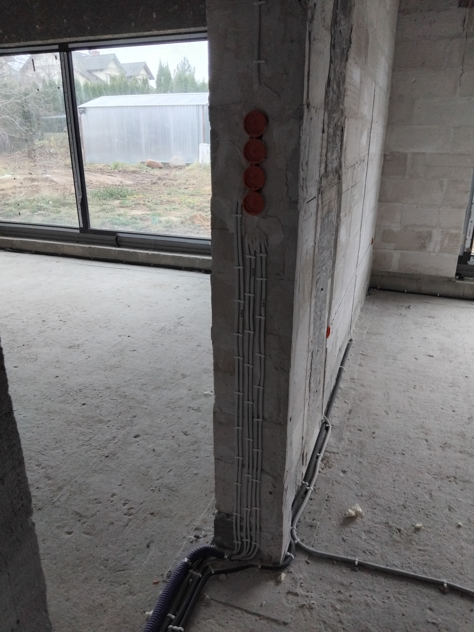 Surowy betonowy filar z pionowo ułożonymi przewodami elektrycznymi i pomarańczowymi puszkami. W tle okno z widokiem na zewnątrz budynku.