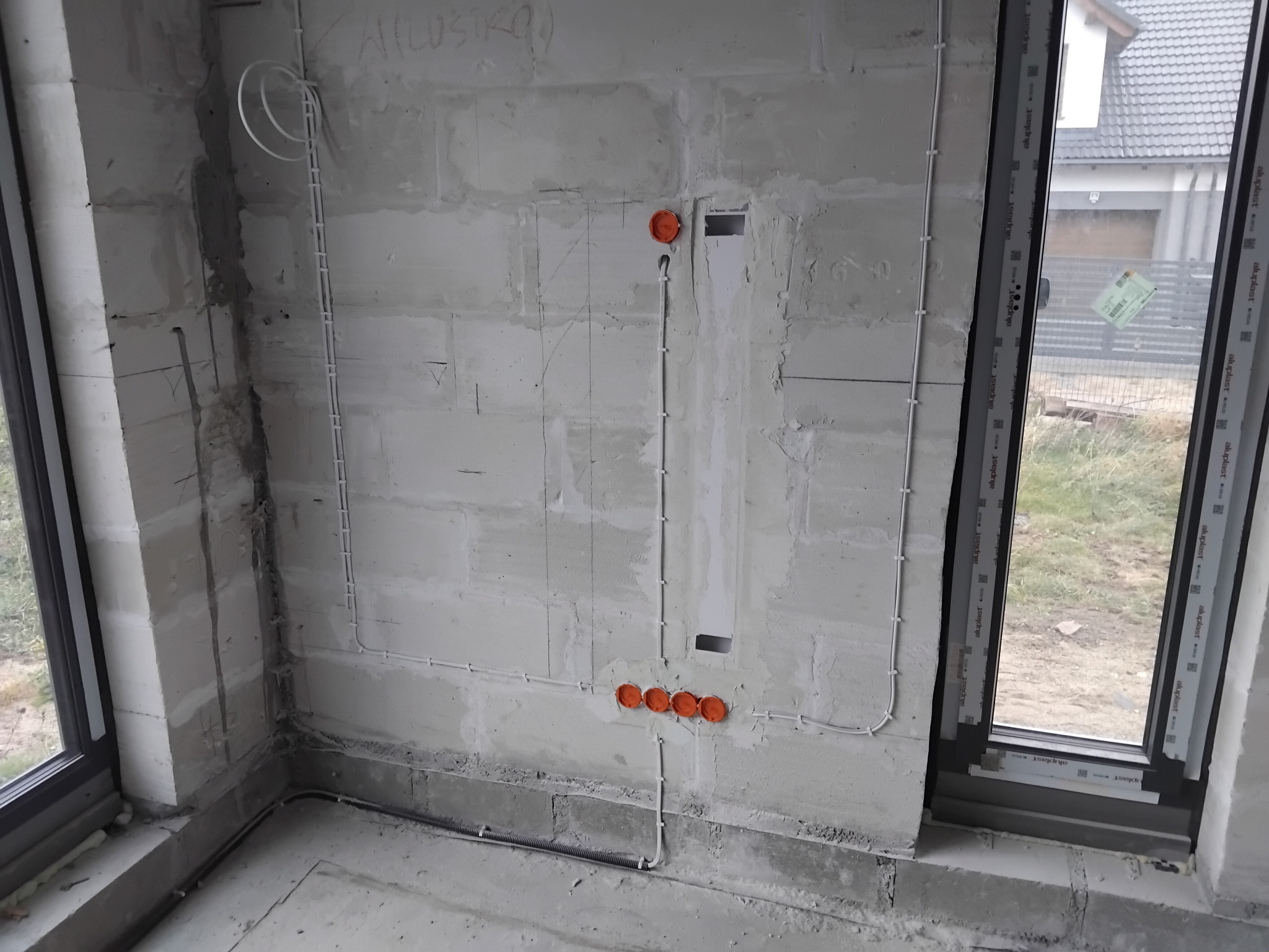 Wewnętrzna instalacja elektryczna w surowym stanie: białe kable prowadzone po ścianie z betonu komórkowego, pomarańczowe puszki elektryczne. Widok przez okno na zewnątrz.