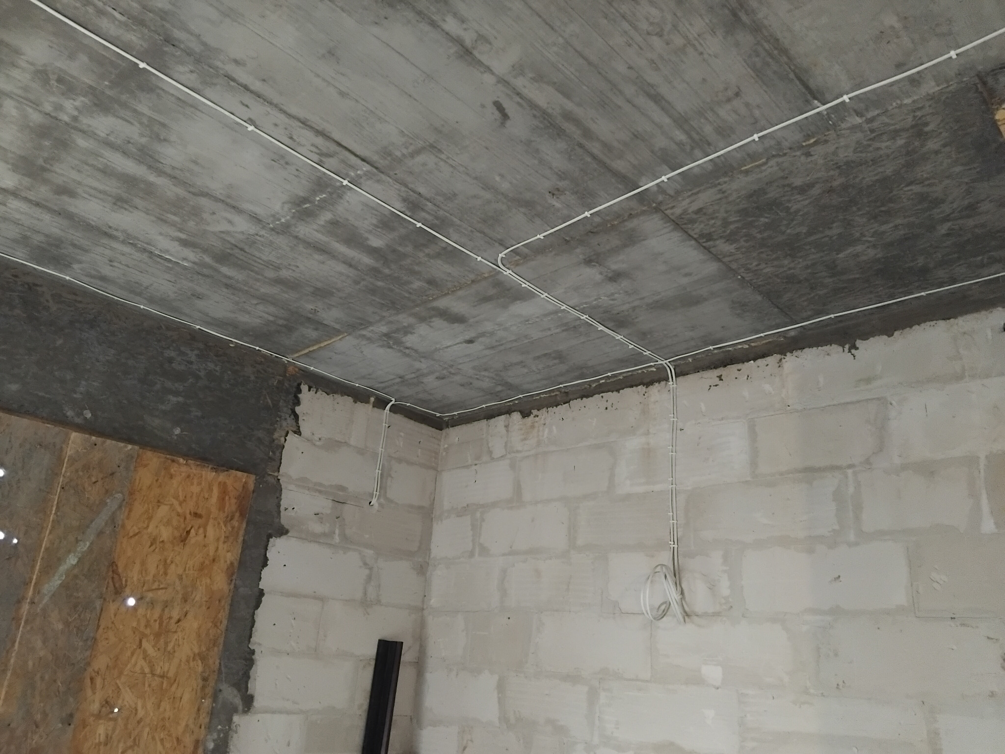 Surowy strop z widoczną, białą instalacją elektryczną w peszlach, prowadzoną po powierzchni betonu. Ściany z białych bloczków, w rogu płyta OSB.