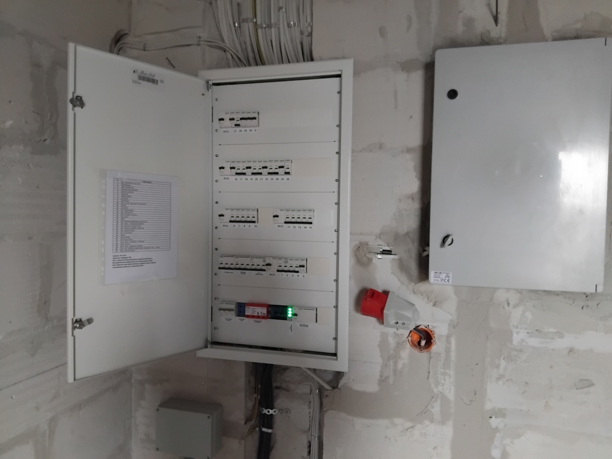 Otwarta skrzynka elektryczna z bezpiecznikami na surowej ścianie. Widoczne kable, gniazdo siłowe i druga, zamknięta skrzynka. Etap prac instalacyjnych.