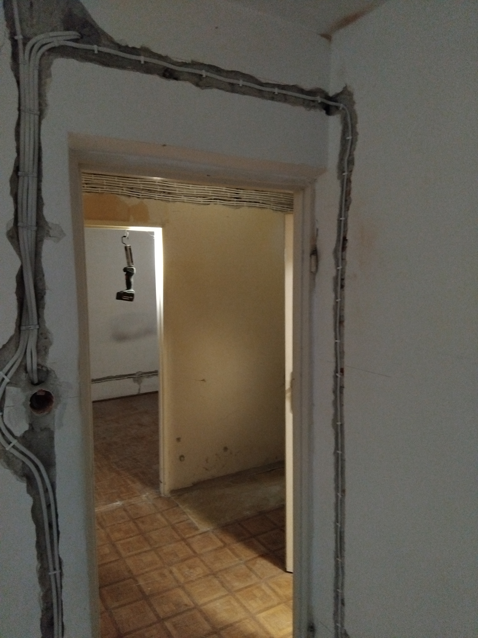 Ściana z bruzdami i przewodami elektrycznymi w trakcie montażu, widok na sąsiednie pomieszczenie. W tle wiertarka wisząca na haku.