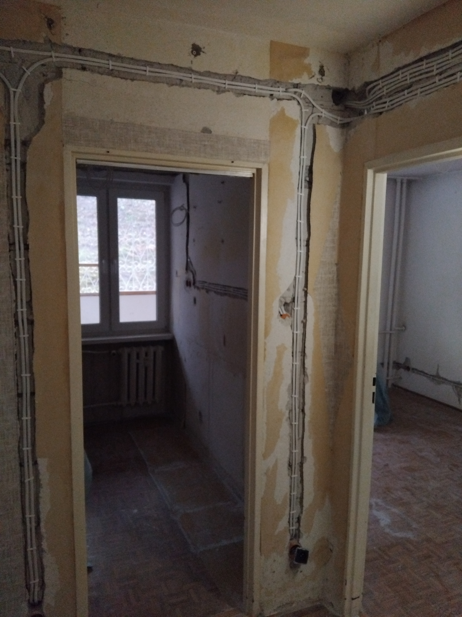 Białe kable elektryczne ułożone wzdłuż ścian w trakcie remontu mieszkania. Widoczne puszki instalacyjne i fragment okna. Nieotynkowane ściany.