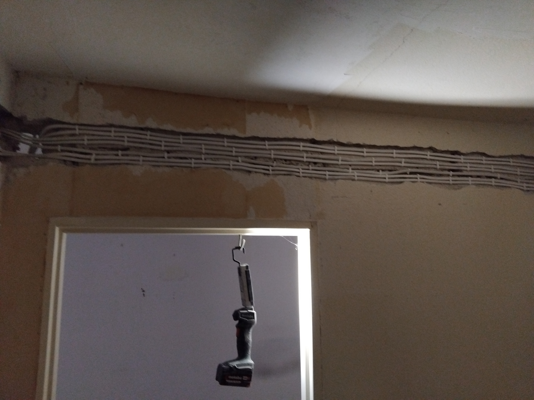 Rzędy białych kabli elektrycznych ułożone w bruździe w ścianie, widoczne narzędzie pracy - lampa warsztatowa. Ściana w trakcie remontu, z widocznymi fragmentami starej farby.