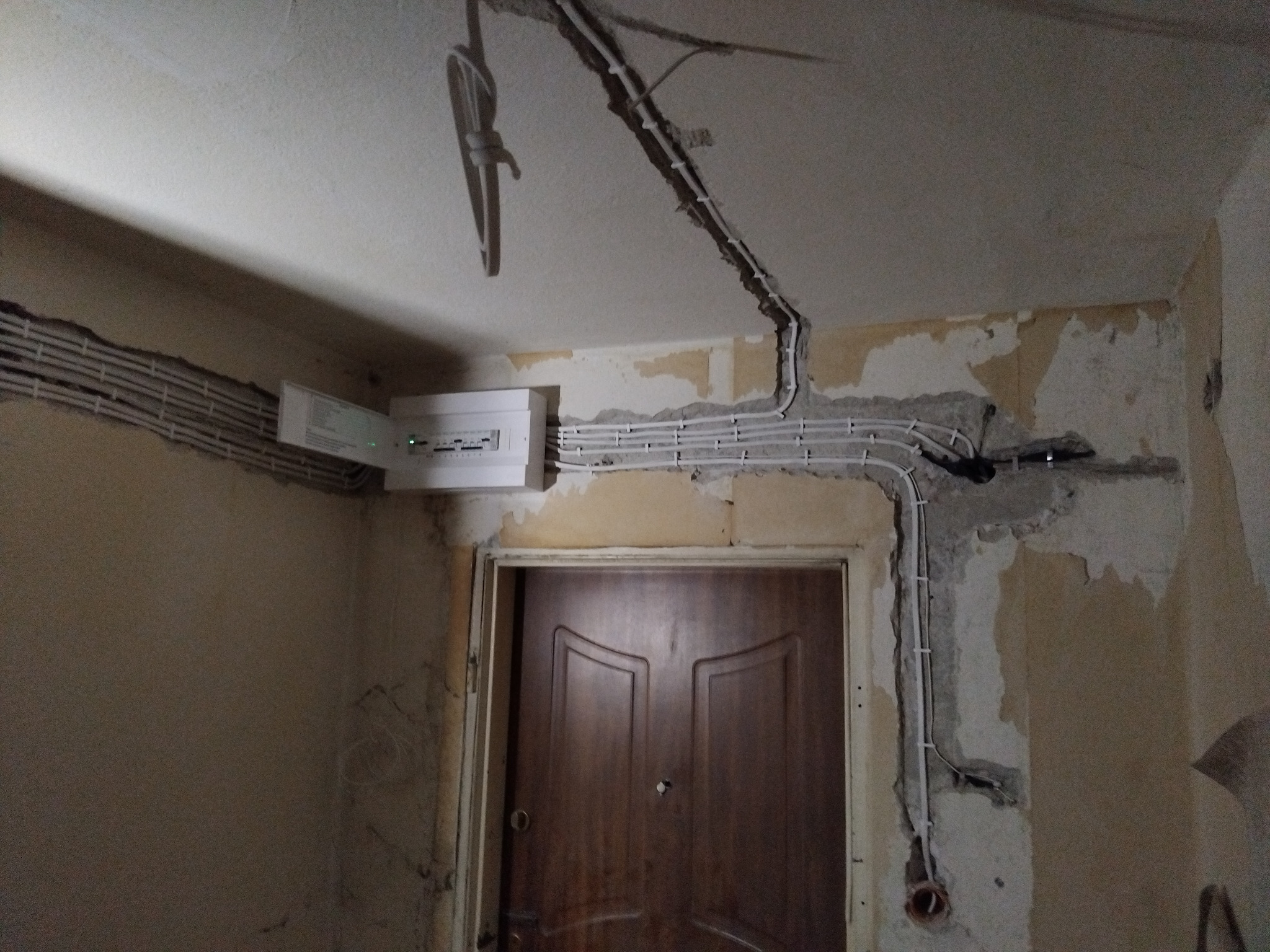 Rozprowadzenie przewodów elektrycznych w bruzdach ściennych, widoczna skrzynka rozdzielcza z otwartymi drzwiczkami i wystające kable. Ściany w trakcie remontu, widoczne fragmenty starej tapety.