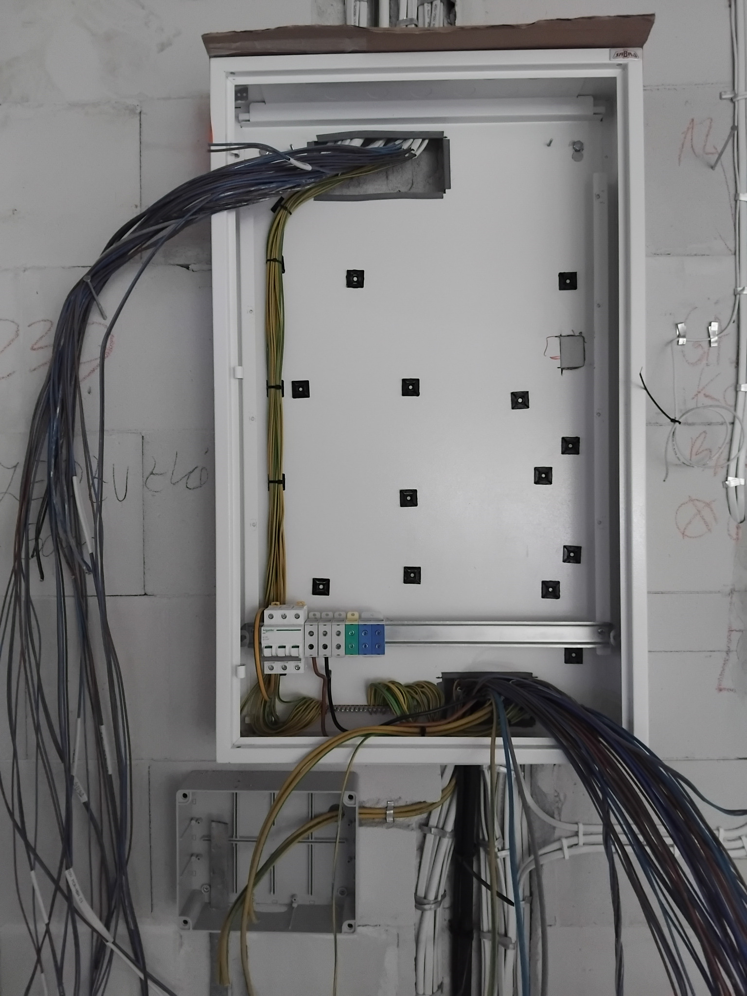 Rozdzielnica elektryczna w trakcie montażu, widoczne kable w różnych kolorach, zabezpieczenia i pusta przestrzeń na osprzęt w surowym wnętrzu budynku.