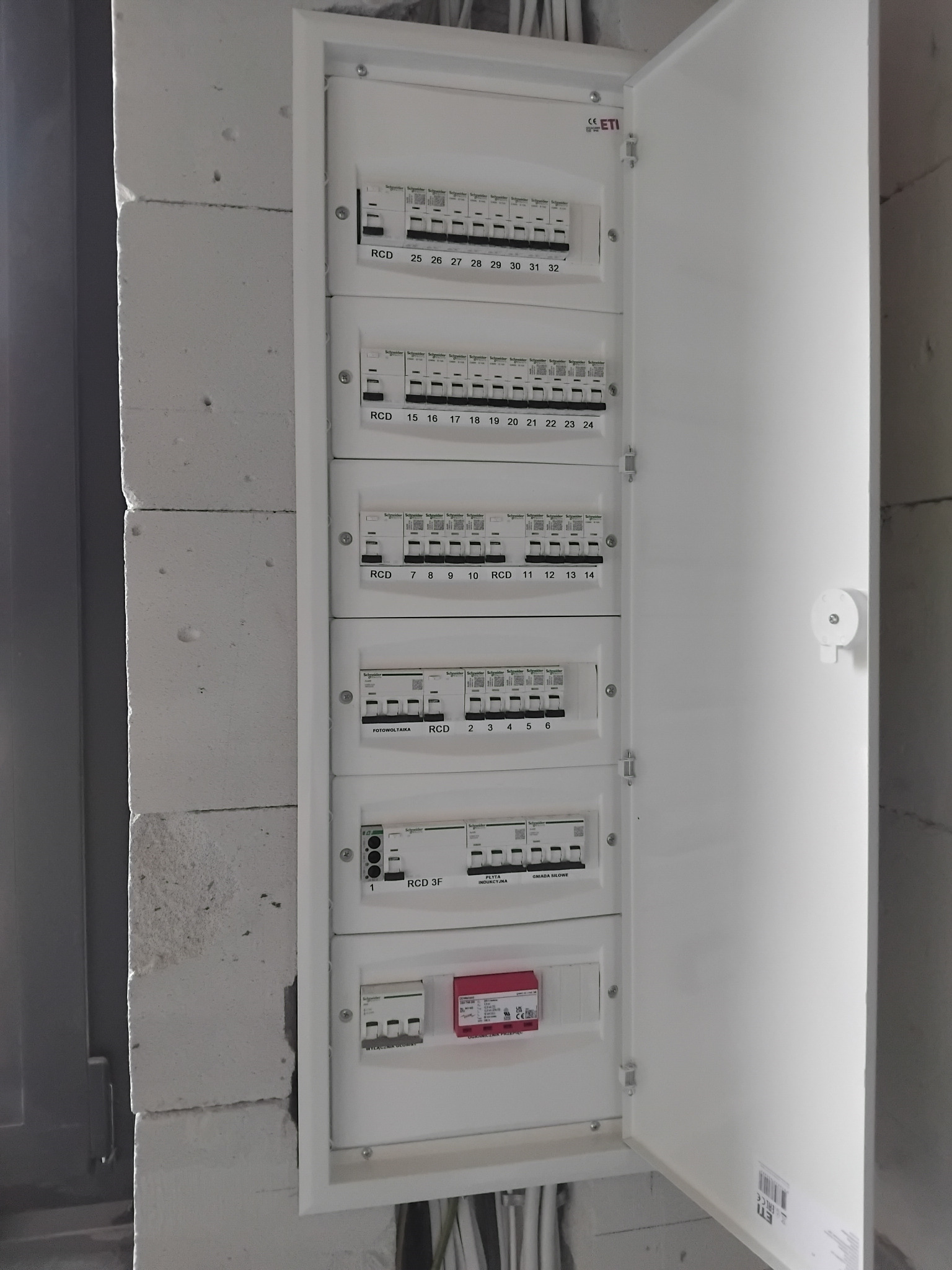 Otwarta, biała skrzynka rozdzielcza z bezpiecznikami i wyłącznikami różnicowoprądowymi, zamontowana na ścianie z białych bloczków. Widoczne kable w dolnej części.
