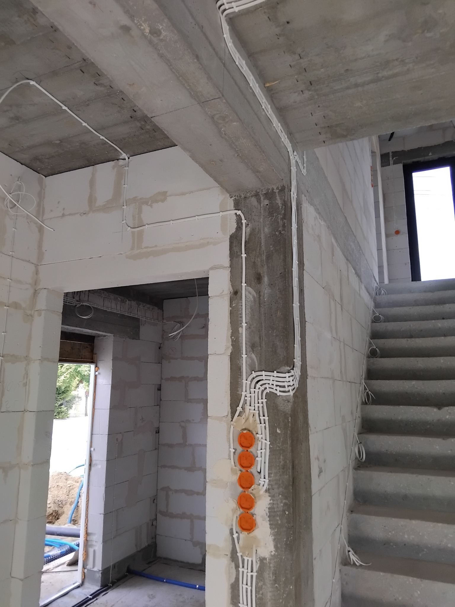 Białe kable elektryczne, pomarańczowe puszki podtynkowe na surowej ścianie z betonu komórkowego wewnątrz budynku w trakcie instalacji. Widoczne schody i otwór drzwiowy.