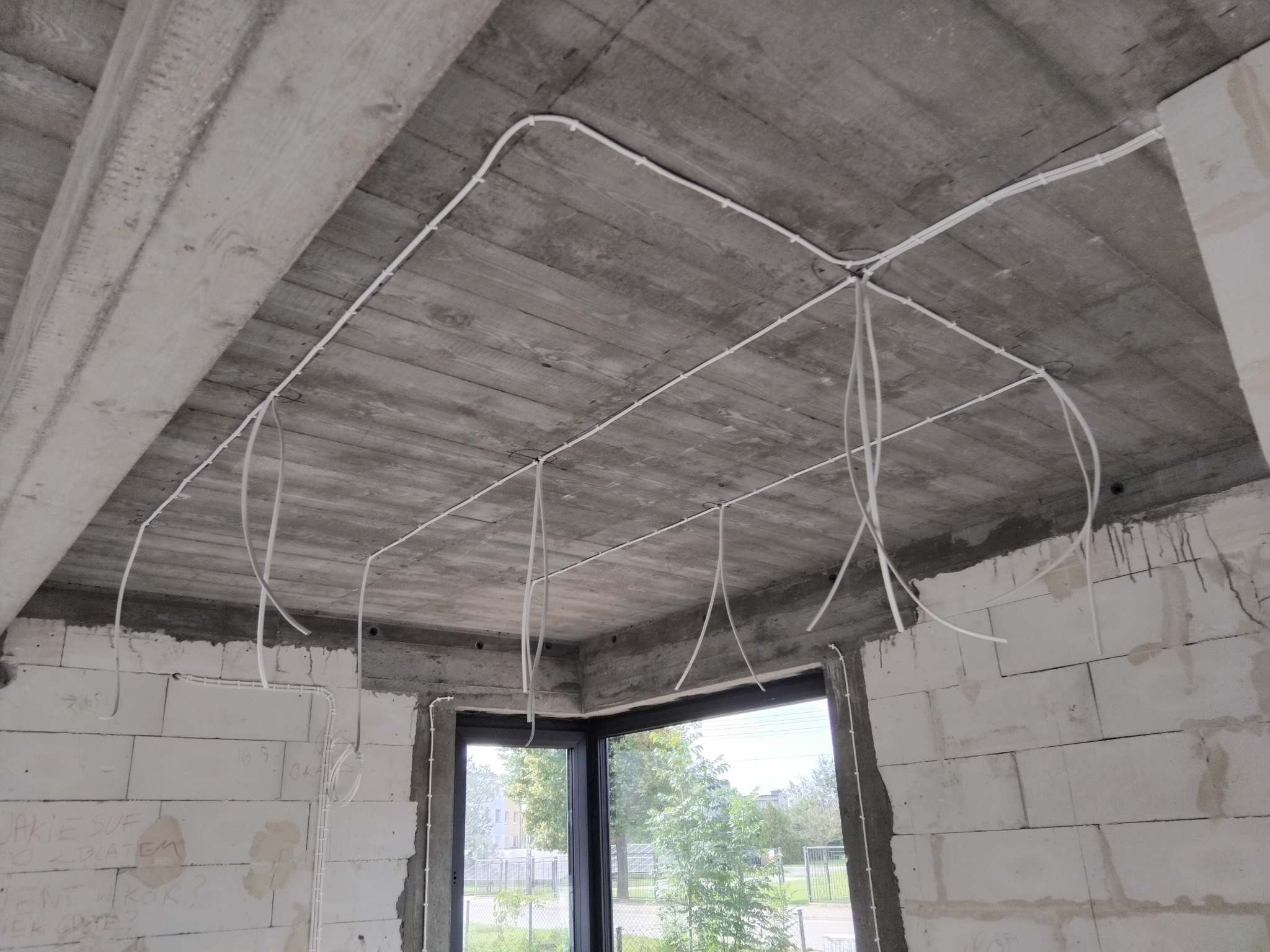 Surowy strop z widoczną, białą instalacją elektryczną w peszlach, zwisającą luźno. W tle okno z widokiem na zieleń. Ściany z białego betonu komórkowego z napisami.