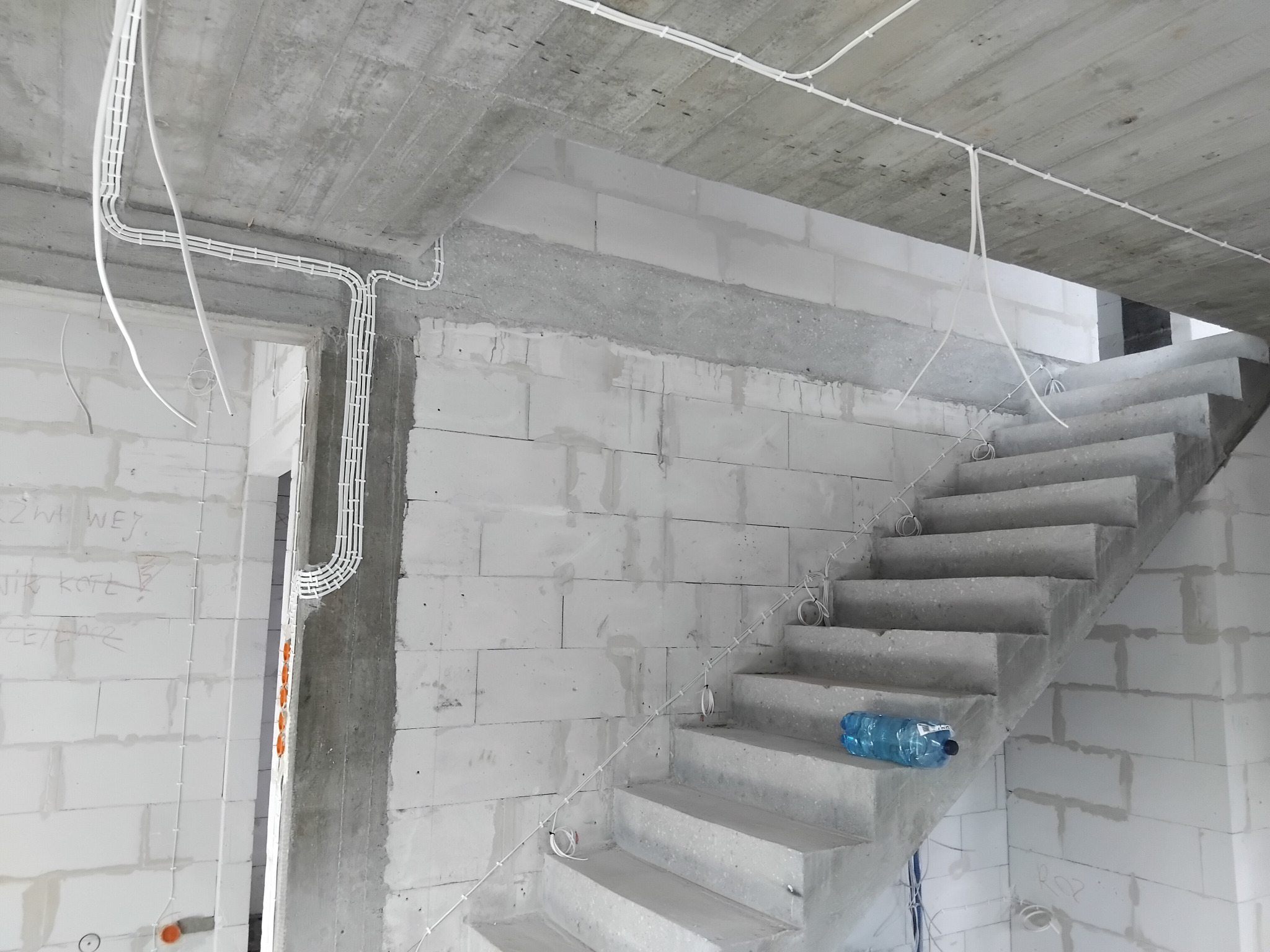 Surowy stan: białe kable elektryczne w plastikowych listwach na ścianach i suficie z betonu komórkowego, surowe betonowe schody z butelką wody na jednym ze stopni.