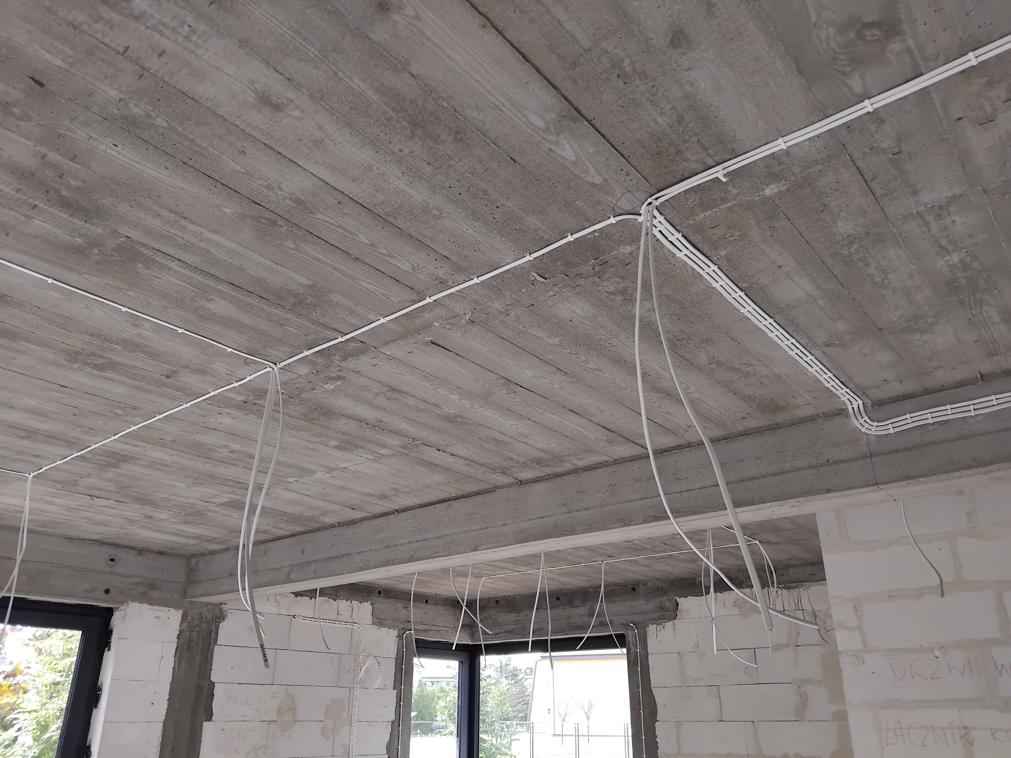 Białe kable elektryczne prowadzone po szorstkim, betonowym suficie w surowym wnętrzu budynku w trakcie budowy. Widoczne okna i ściany z białego bloczka.