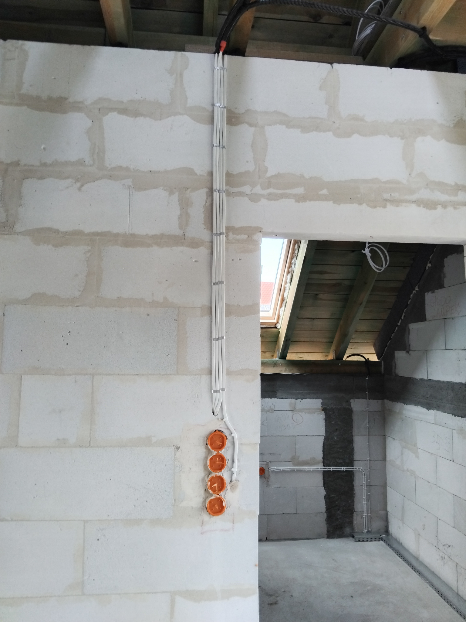Instalacja elektryczna w surowym wnętrzu: białe przewody poprowadzone po ścianie z bloczków, pomarańczowe puszki pod gniazdka, widok na drewnianą konstrukcję dachu.