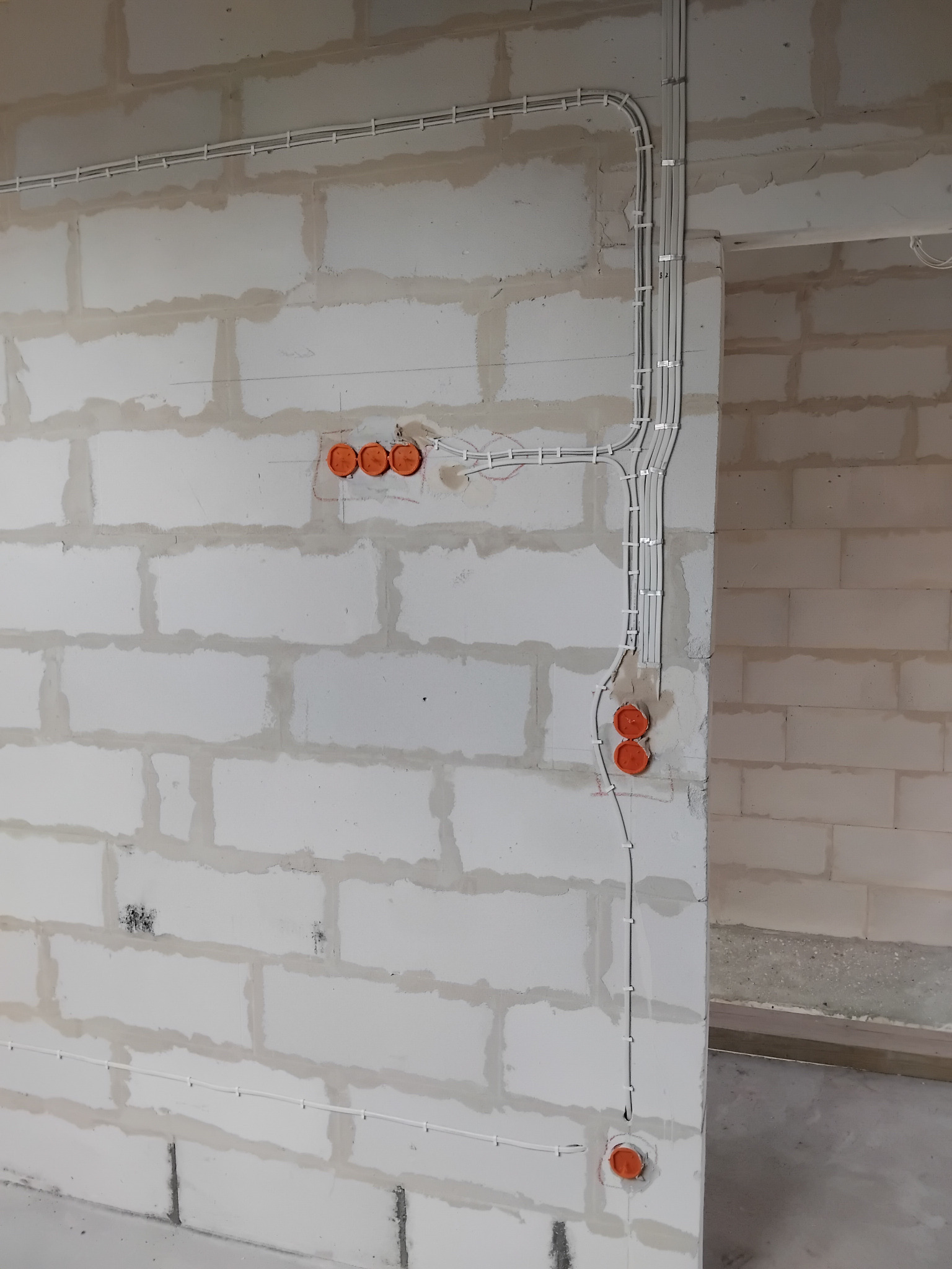 Biała ściana z betonu komórkowego z zamontowanymi, białymi przewodami elektrycznymi w listwach i pomarańczowymi puszkami instalacyjnymi pod gniazdka.