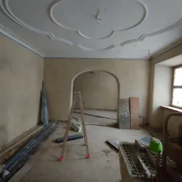 Wnętrze w trakcie tynkowania maszynowego, widoczna drabina, agregat tynkarski, listwy oraz ozdobny sufit z gładzią