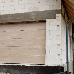 Jasnobrązowa segmentowa brama garażowa z poziomymi panelami o fakturze drewna, zamontowana w surowym murze z bloczków betonowych na etapie budowy domu.