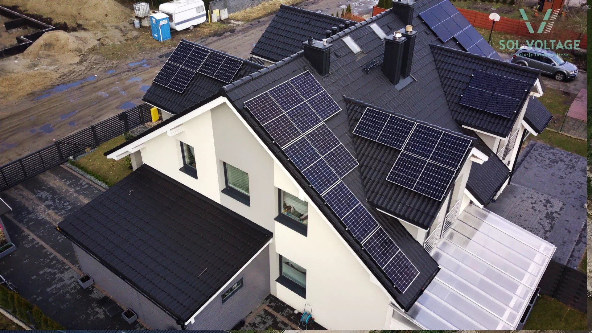 Widok z góry na dom z panelami słonecznymi na dachu pokrytym czarną dachówką, z widocznym logo firmy instalującej panele, otoczenie domu częściowo w trakcie prac ziemnych.