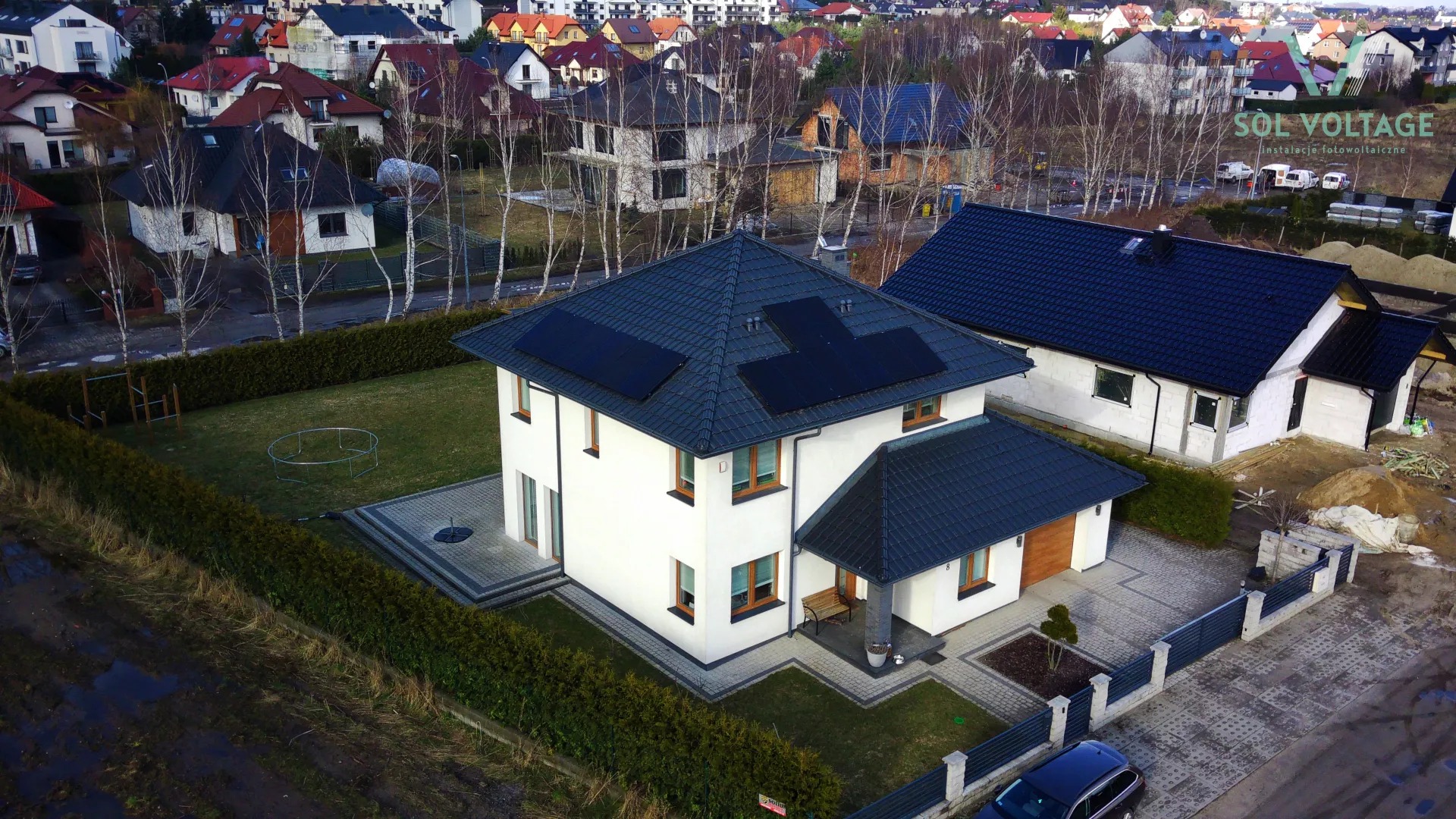 Nowoczesny dom jednorodzinny z panelami słonecznymi na dachu, widok z góry, w tle zabudowa osiedlowa i logo firmy instalującej.
