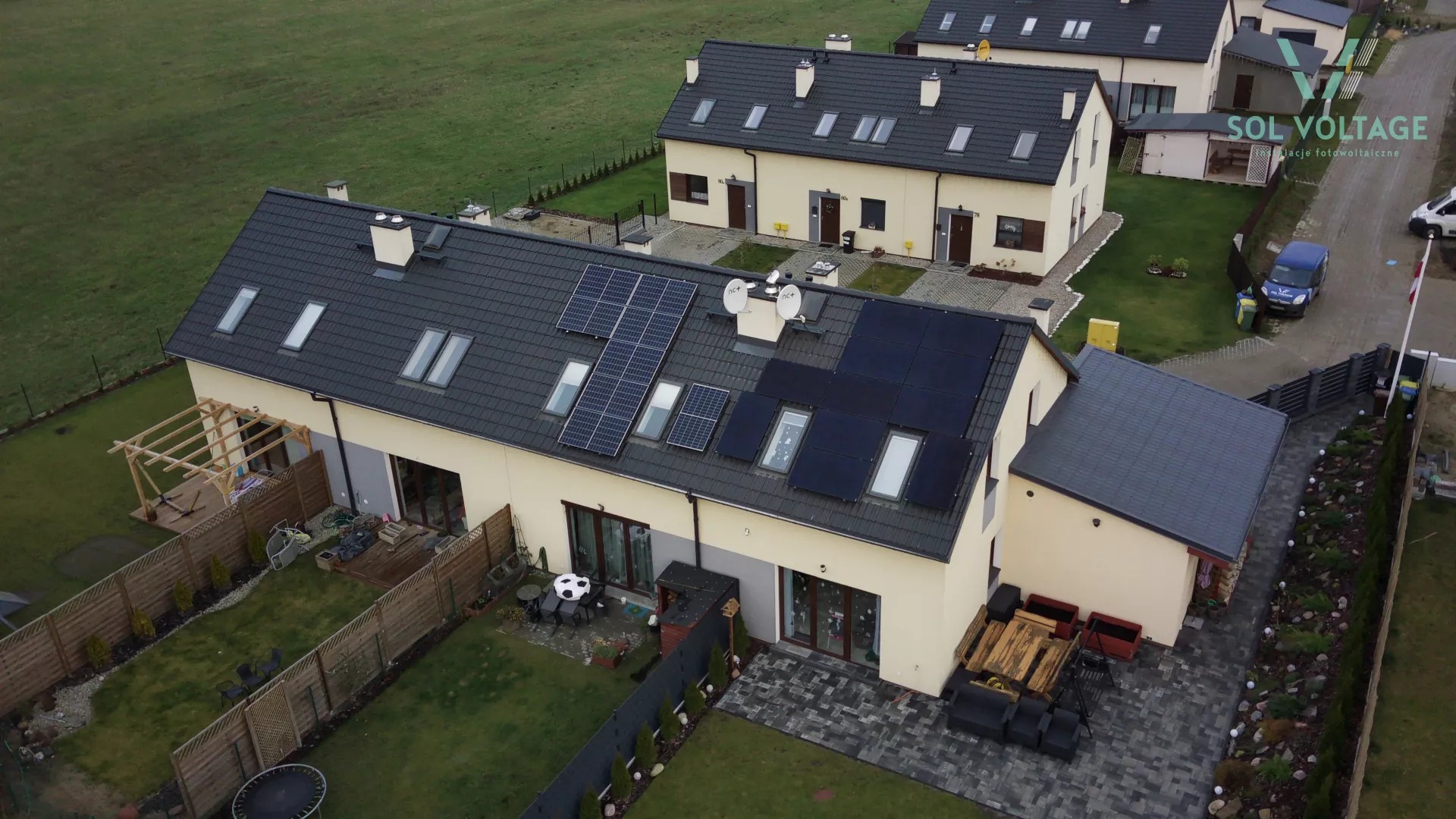 Widok z góry na domy szeregowe z panelami słonecznymi na dachu, różna konfiguracja paneli, logo firmy na zdjęciu, zadbane ogrody.