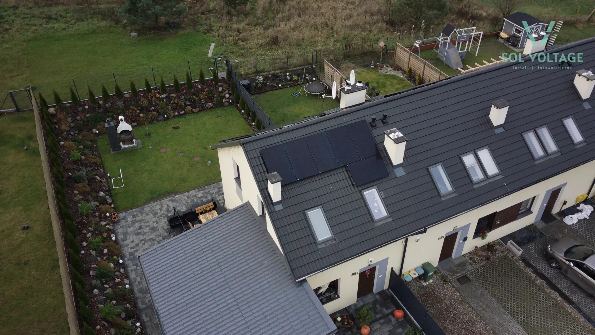 Widok z góry na szeregowy dom z ciemną dachówką, na której zamontowane są cztery czarne panele, z zadbanym ogrodem z grillem i trampoliną oraz logo firmy instalującej panele w prawym górnym rogu.