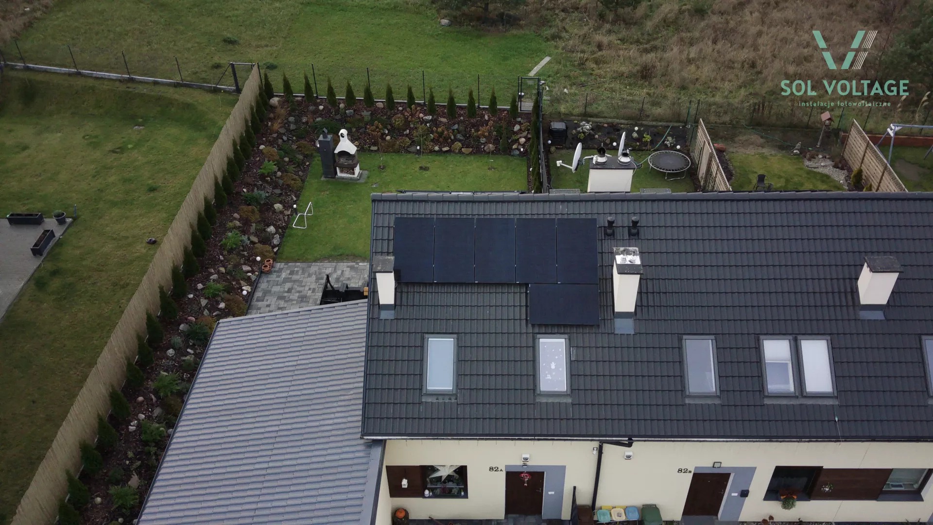 Widok z góry na dom z zamontowanymi panelami słonecznymi na dachu, ogród z grillem i trampoliną w tle, logo firmy instalującej panele w prawym górnym rogu.