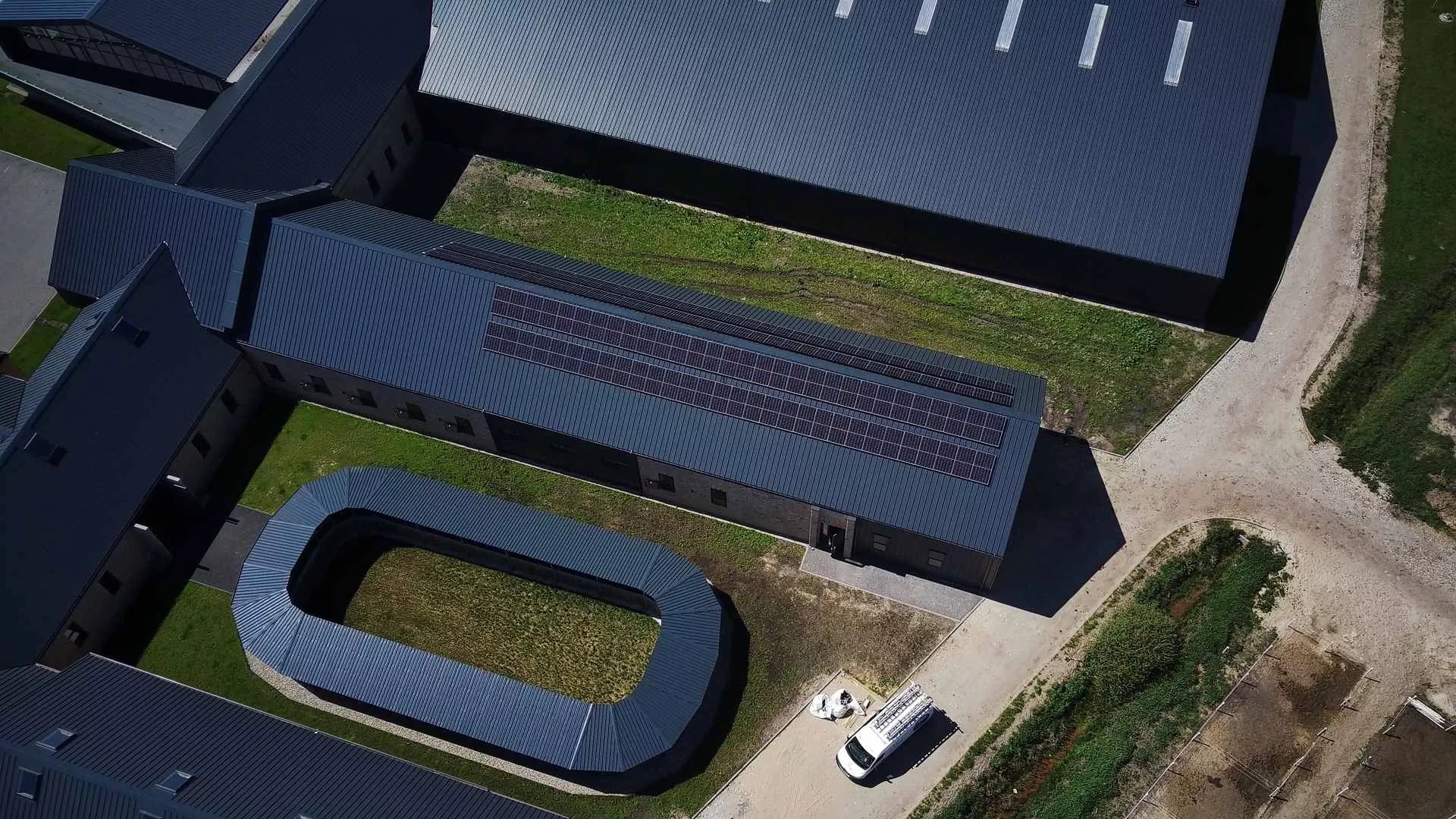Widok z drona na budynek z panelami słonecznymi na dachu, obok zaparkowany biały samochód dostawczy z drabiną na dachu i sprzętem obok, otoczony zielenią i innymi budynkami z ciemnymi dachami.
