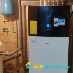 Zamontowana jednostka wewnętrzna pompy ciepła Daikin z widocznymi przyłączami hydraulicznymi i zbiornikiem wyrównawczym na tle ściany z boazerią i widocznym logo firmy A2 Energy.