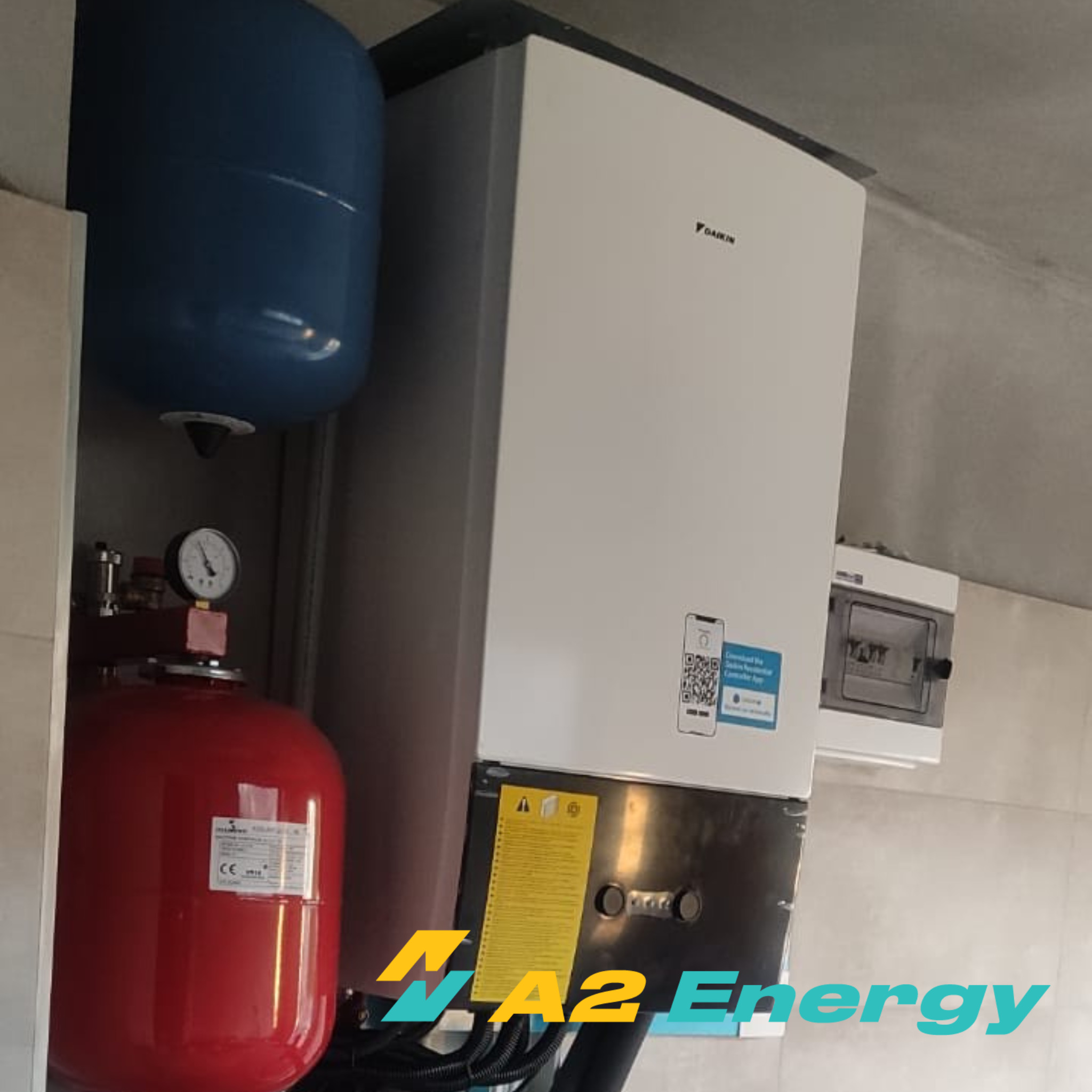 Biała jednostka Daikin z niebieskim zbiornikiem wyrównawczym, czerwonym naczyniem przeponowym i skrzynką elektryczną na szarej ścianie.