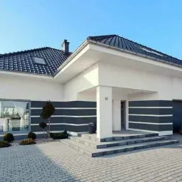 Nowoczesny dom jednorodzinny z białymi ścianami i grafitowymi akcentami, widok z zewnątrz na elewację z oknem i wejściem, kostka brukowa na podjeździe.