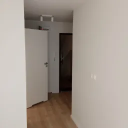 Wnętrze korytarza z białymi ścianami, drewnianą podłogą, otwartymi białymi drzwiami i widoczną instalacją elektryczną pod sufitem.