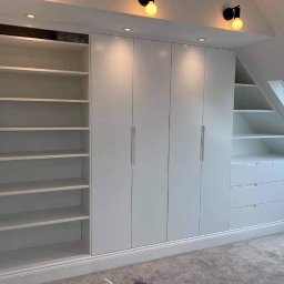 Wood-Pol - Biała zabudowa meblowa pod skosem, z szafami trzydrzwiowymi, otwartymi półkami i szufladami, oświetlona punktowo, z widocznym oknem dachowym.