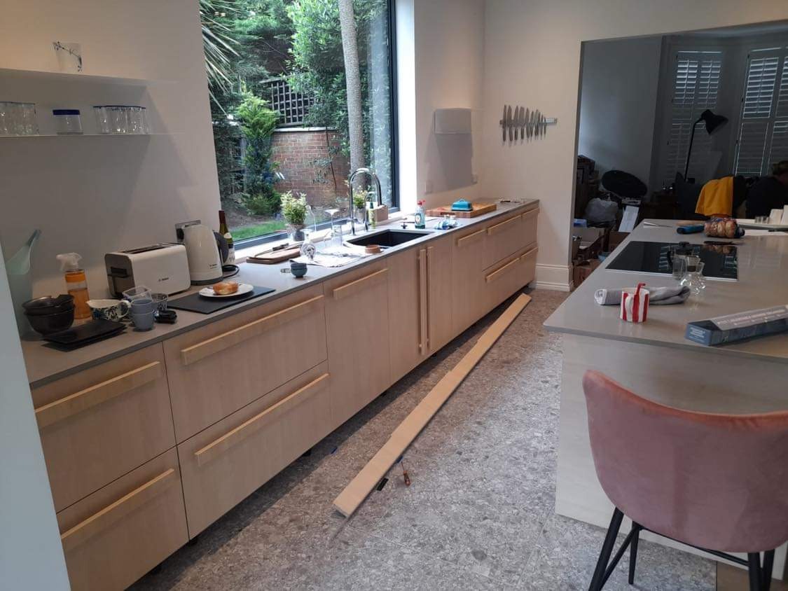 Jasna, nowoczesna kuchnia z drewnianymi frontami szafek i szarym blatem, widok na zewnątrz przez duże okno, w tle wyspa kuchenna z różowym krzesłem, na podłodze listwa przypodłogowa w trakcie montażu.