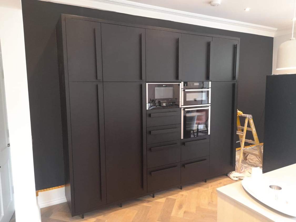 Ciemna zabudowa kuchenna z piekarnikami i mikrofalówką, minimalistyczne fronty szafek z podłużnymi uchwytami, na podłodze parkiet w jodełkę, drabina malarska w tle.