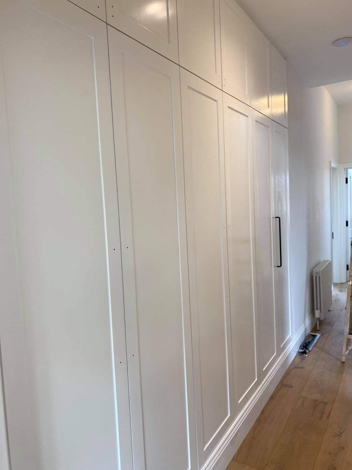 Wysoka, biała zabudowa meblowa z frezowanymi frontami, sięgająca sufitu, z długimi, czarnymi uchwytami, widziana z perspektywy pod kątem w korytarzu na tle grzejnika.