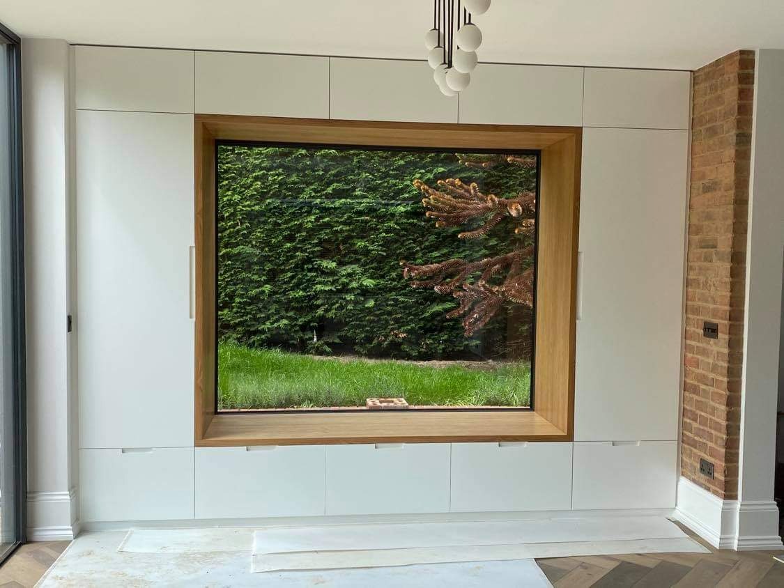 Biała zabudowa meblowa z drewnianą ramą okienną, widok na zielony ogród, minimalistyczny design, parkiet w jodełkę zabezpieczony kartonem.