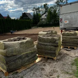 Palety z trawą w rolkach, zabezpieczone siatką, stoją obok samochodu dostawczego na przygotowanym podłożu, w tle żywopłot i domy.