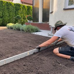 SADZIMYTO - Grzegorz Malinowski - Pracownik wyrównuje ziemię listwą aluminiową przed domem z zielonym żywopłotem. Przygotowanie terenu pod przyszły trawnik lub rabatę.