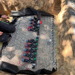 SADZIMYTO - Grzegorz Malinowski - Instalacja systemu nawadniania w wykopie, widoczne rury, elektrozawory, przewody, geowłóknina i warstwa drenażowa z kamienia. Ujęcie z góry, w trakcie montażu.