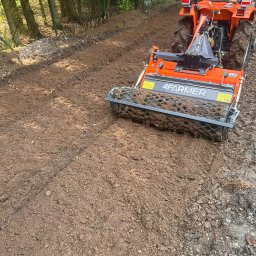 SADZIMYTO - Grzegorz Malinowski - Pomarańczowy traktor 4Farmer przygotowuje glebę pod przyszły ogród, wyrównując ziemię na działce. Widoczne świeże ślady opon i spulchniona ziemia.