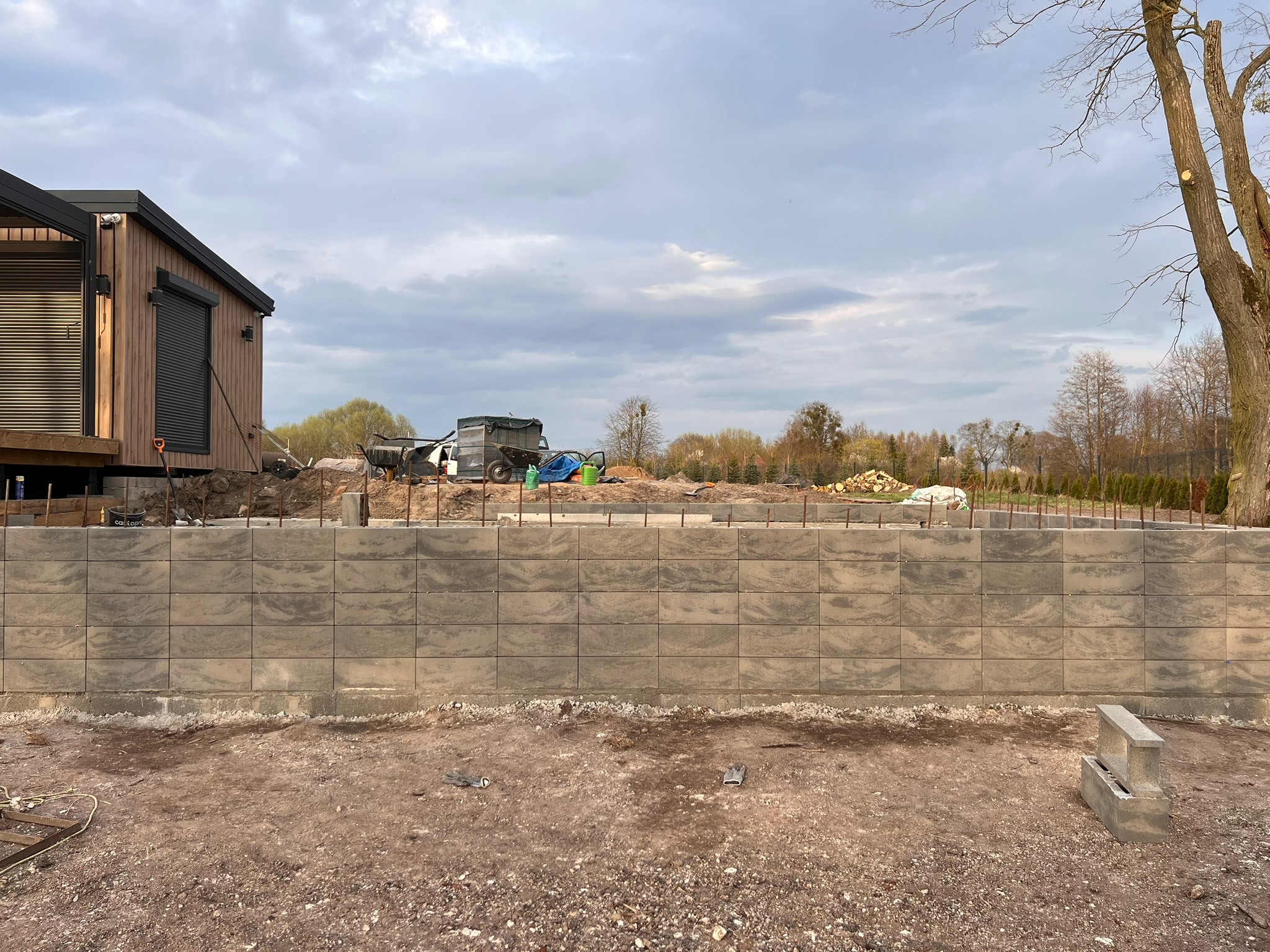 Wznoszenie muru oporowego z szarych, betonowych bloczków przy nowoczesnym domu. Widoczne zbrojenia i przygotowany teren pod przyszły ogród. Pochmurne niebo w tle.