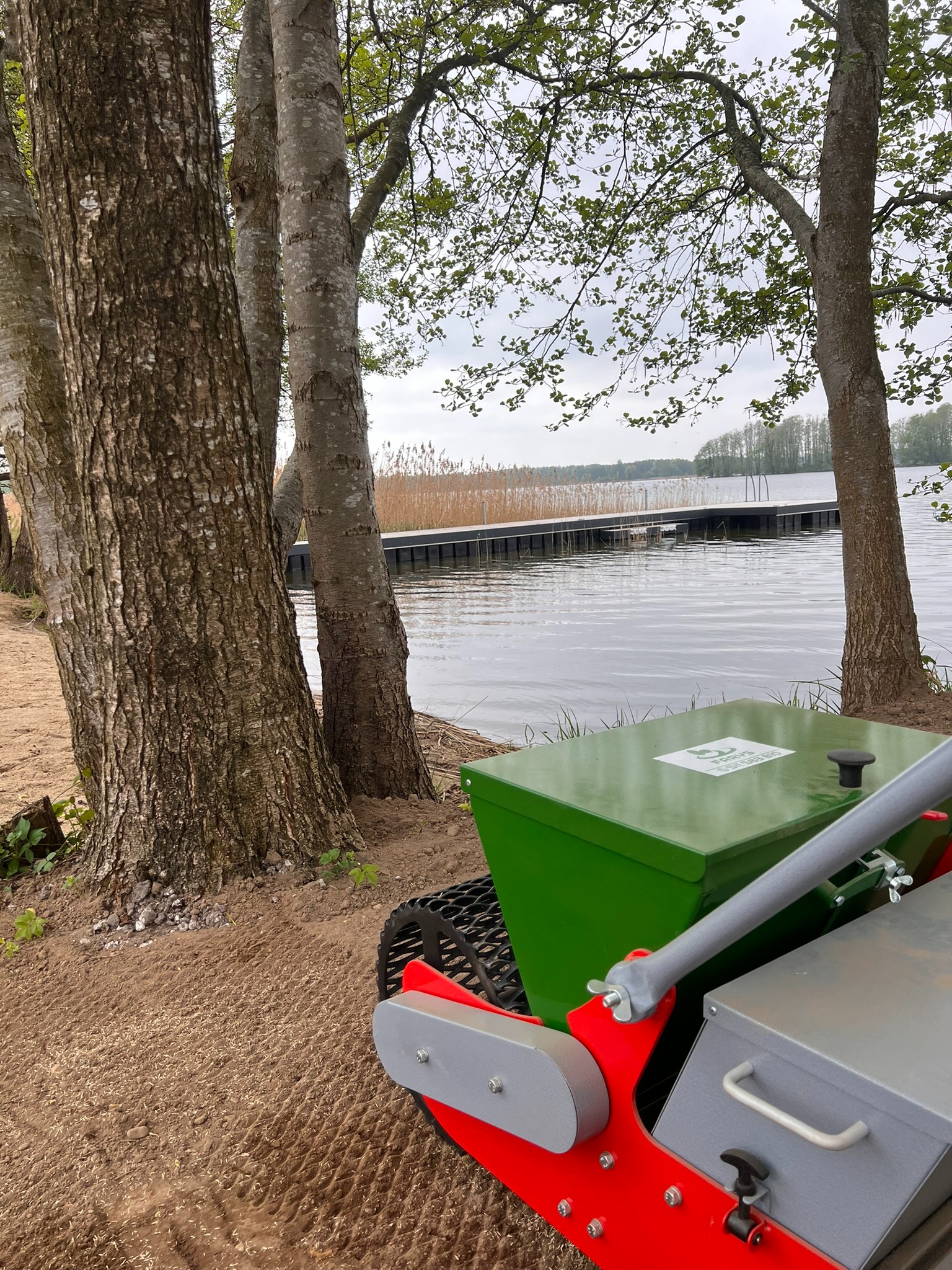 Specjalistyczna maszyna do siewu trawy na piaszczystym terenie przy jeziorze, widoczne ślady po siewie. W tle pomost i trzciny.