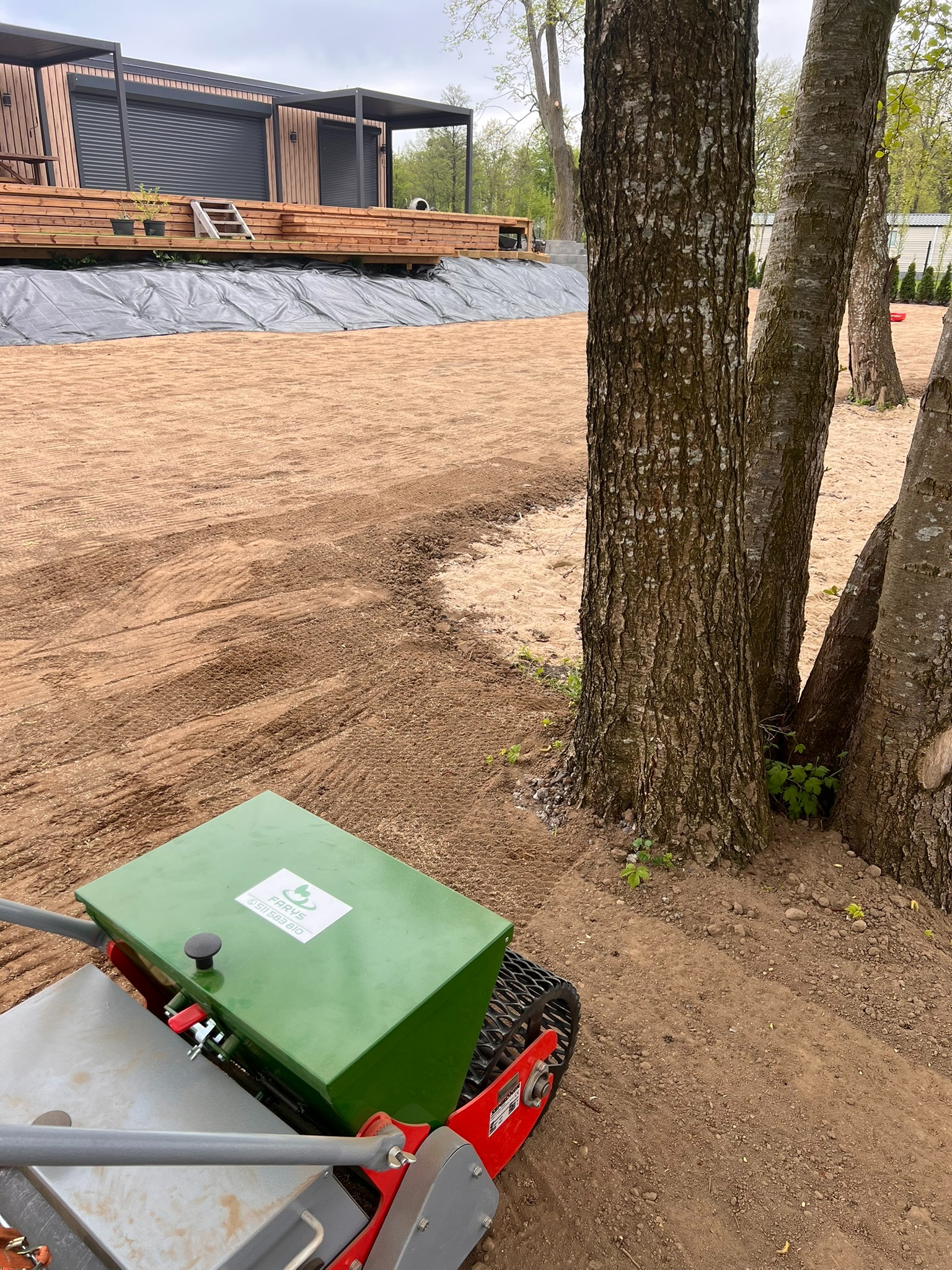 Przygotowanie terenu pod trawnik: wyrównana ziemia, maszyna do siewu trawy, widoczne drzewa i fragment domu z tarasem w tle. Etap prac ziemnych.